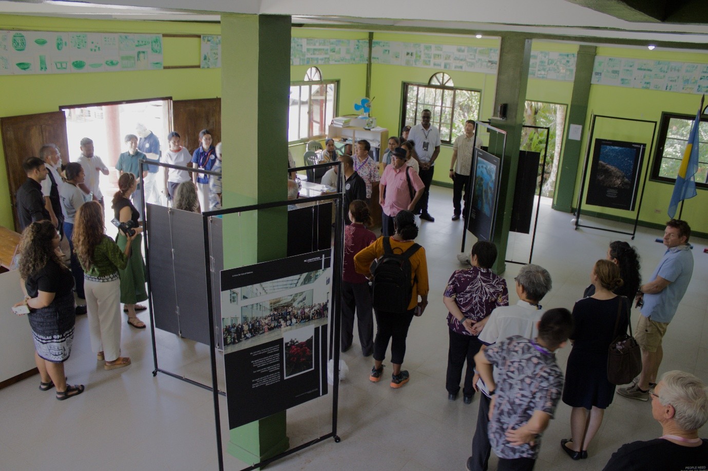 A photo exhibition set up in a large room with painted green walls. People walk about looking at the photos displayed on boards